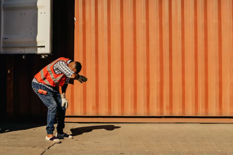 Worker in safety vest leaning over expressing heat exhaustion standing next to orange shipping container.